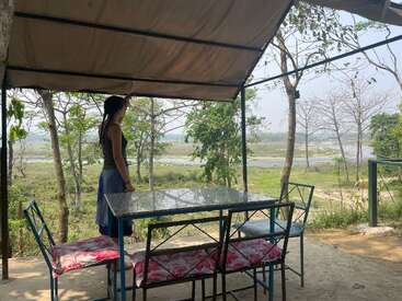 A woman stands under a canopy, beside a glass table with four chairs, gazing at a scenic, green riverside landscape with trees and clear, sunny skies.