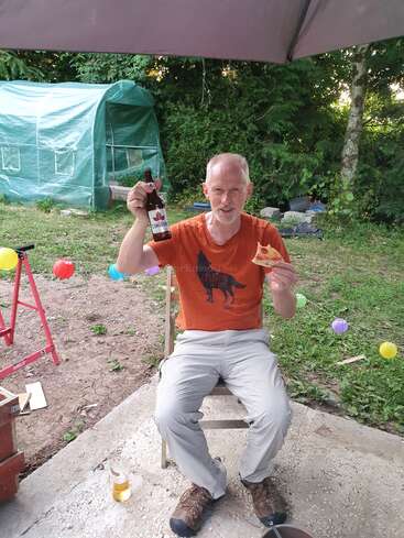A man sits outdoors under a canopy, holding a slice of pizza and a bottle of Canadian beer, surrounded by colorful decorations and a green tent.