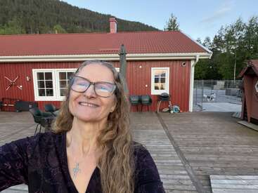 A smiling woman with long hair and glasses takes a selfie on a wooden deck. Behind her is a red house, outdoor furniture, and trees.