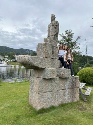 Three girls pose on a large stone sculpture resembling a human figure by a scenic lakeside, with green grass, trees, mountains, and cloudy skies in the background.