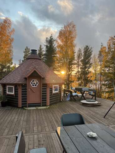 A cozy wooden cabin sits on a spacious deck, surrounded by autumn trees. The sun sets behind, casting a golden glow, with outdoor furniture nearby. Peaceful atmosphere.
