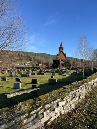 A wooden church with a tall steeple stands beyond a graveyard, surrounded by stone walls, bare trees, green grass, and hills under a clear blue sky.