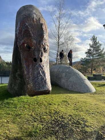 A large, realistic sculpture of a sea serpent's head and body sits on green grass. Two people stand on its back, surrounded by trees and sky.