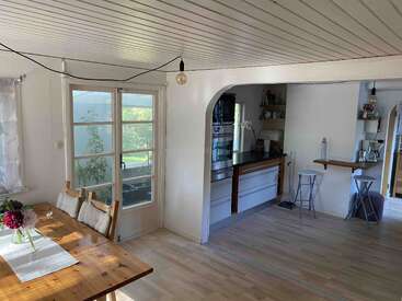 This image shows a bright, cozy kitchen and dining area with wooden furniture, light-colored walls, a glass door, and minimalistic decor. Natural light fills the space.