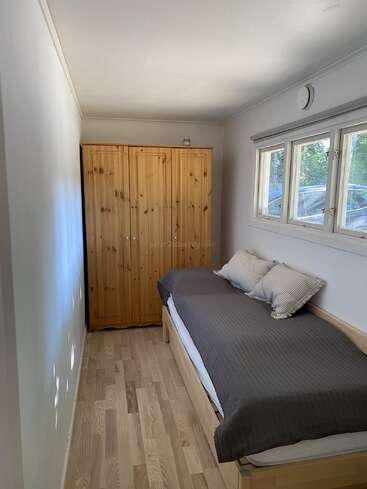 This image shows a small, narrow bedroom with light wooden floors, a single bed with gray bedding, pillows, a wooden wardrobe, and three windows providing daylight.