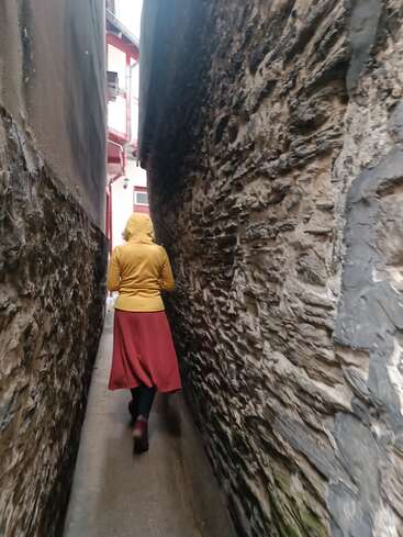 Eine Person in einem gelben Kapuzenpulli und einem roten Rock geht durch eine sehr schmale Gasse zwischen Steinmauern, an deren Ende Licht aufleuchtet.