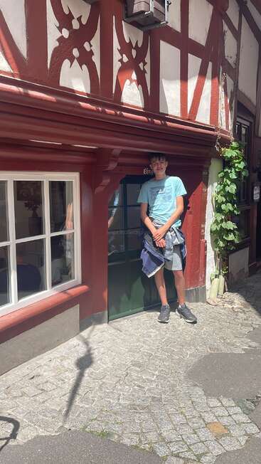 Ein großer junger Mann steht an einem sonnigen Tag an der winzigen Tür eines charmanten, traditionellen Fachwerkhauses, lächelt und genießt die malerische Straßenszene.