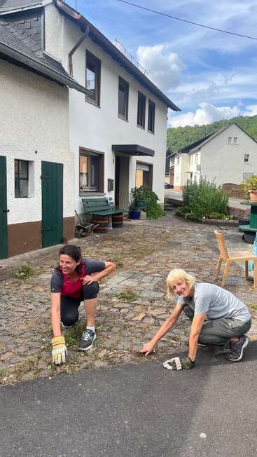 Zwei Frauen hocken vor einem Haus und zupfen Unkraut zwischen den Pflastersteinen heraus. Sie lächeln, tragen Handschuhe und genießen einen sonnigen Tag in einer malerischen Gegend.
