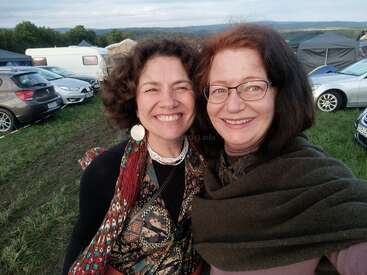 Zwei Frauen, die bei einer Veranstaltung im Freien lächeln, umgeben von Autos und Zelten auf einer Wiese, genießen einen schönen Tag mit malerischen Hügeln im Hintergrund.