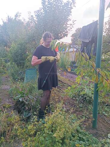 Eine Person, die ein schwarzes Kleid, Handschuhe und Stiefel trägt, arbeitet in einem üppigen Garten. Grüne Pflanzen und Bäume umgeben sie, und in der Nähe hängen Kleider zum Trocknen.