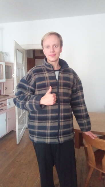 Eine Person steht in einem Haus und trägt eine blau-beige karierte Jacke. Sie lächelt, zeigt den Daumen nach oben und steht neben einem Holztisch und Stühlen.