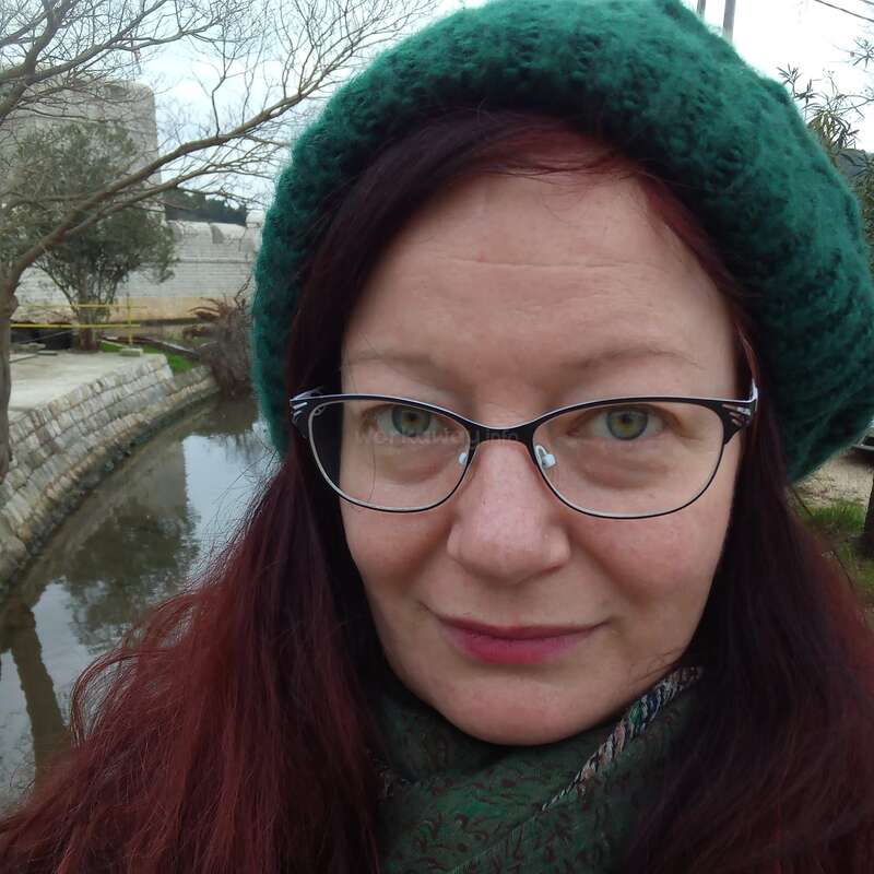 Das Bild zeigt eine Frau mit langen roten Haaren, die eine grüne Baskenmütze und einen Schal trägt und vor einem kleinen Teich und einer Steinmauer steht, mit Bäumen und Gebäuden im Hintergrund.