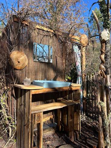 This image shows a rustic outdoor sink setup with wooden elements, a blue basin, a mirror, natural decor, and sunlight shining through trees against a clear sky.
