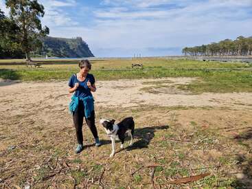 Eine Frau steht draußen mit einem schwarz-weißen Hund auf einer sandigen, grasbewachsenen Wiese. Bäume, Bänke, ferne Hügel und ein strahlend blauer Himmel bilden eine friedliche Szenerie.
