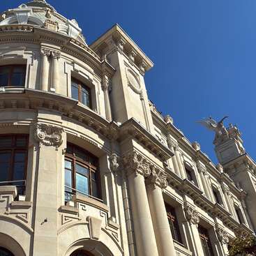 This image shows an ornate, classical building with columns, detailed carvings, arched windows, and a winged statue on the roof, all under a clear blue sky.