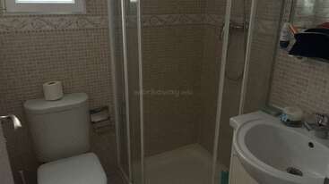 Cette image montre une petite salle de bain avec des murs carrelés de couleur beige, une douche avec des portes en verre, des toilettes, un lavabo et divers articles de toilette soigneusement rangés.