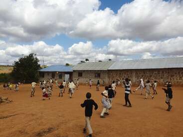 Children are playing energetically in a schoolyard with red soil, next to stone buildings, under a bright sky filled with fluffy white clouds. Joyful atmosphere prevails.