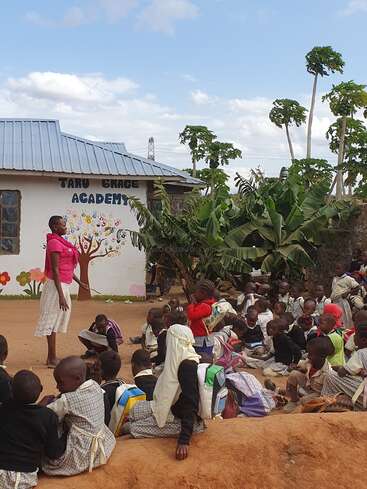 A teacher stands outside instructing a large group of children sitting on the ground near a building labeled “Taru Grace Academy,” surrounded by greenery and trees.