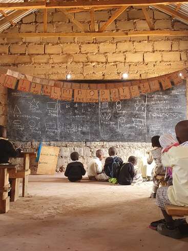 Children sit on wooden benches and the floor of a rustic classroom, facing a large chalkboard filled with lessons; hand-drawn letters hang overhead.