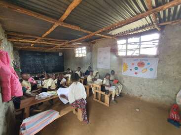 A group of children sit on wooden benches in a rustic classroom, learning with charts on the wall and a blackboard, sunlight streaming through windows.
