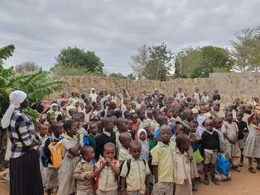 A large group of schoolchildren, most wearing uniforms, stand outdoors in front of a stone wall. Some carry backpacks. A woman stands to the left, watching.