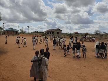 Children in school uniforms play and gather on a red dirt playground under partly cloudy skies, with rural buildings in the background and open landscape.