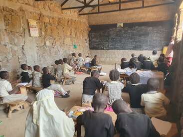 Children are seated on small wooden benches in a rustic classroom with stone walls, attentively facing a large chalkboard, engaged in learning and writing activities.