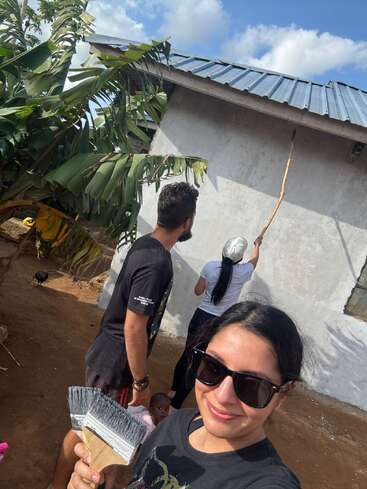 Three people, including a woman smiling in sunglasses, are painting a house's exterior. A small child peeks behind an adult. Tropical plants and blue sky surround them.