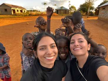 A group of smiling children and two women pose for a cheerful selfie outside on a sunny day, with buildings and trees in the background. Playful atmosphere.