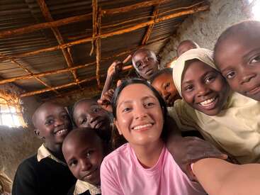 A woman in a pink shirt takes a cheerful selfie with a group of smiling children in a rustic classroom with a wooden roof and stone walls.