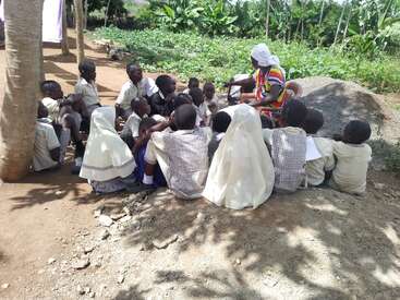 A group of children sit outdoors in a circle, attentively listening to a woman teaching. Lush greenery surrounds them, creating a peaceful, natural classroom setting.