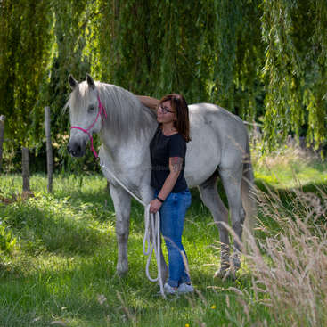 Une femme se tient à côté d'un cheval blanc dans un champ herbeux, la tête du cheval tournée vers la gauche et un licol rose sur son visage.