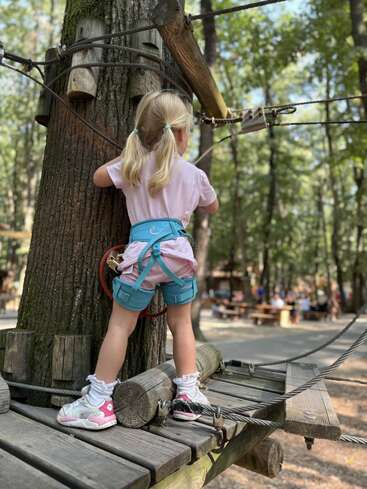 Une jeune fille aux cheveux blonds coiffés en nattes escalade un parcours de cordes en plein air, vêtue d'un harnais de sécurité et de baskets, au milieu des arbres d'un parc.