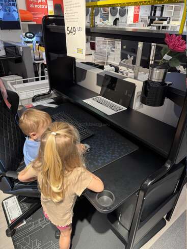 Deux jeunes enfants interagissent avec une installation informatique dans un magasin IKEA, explorant un bureau et une chaise de jeu noirs étiquetés au prix de 549 LEI.
