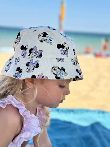 Une jeune fille portant un chapeau de soleil Minnie Mouse et une robe à volants est assise sur une couverture bleue à la plage, avec la mer et le ciel en arrière-plan.