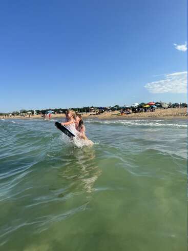 Un enfant jouant joyeusement dans l'eau près d'une plage de sable, tenant une planche de surf, avec un ciel bleu clair et des parasols colorés en arrière-plan.