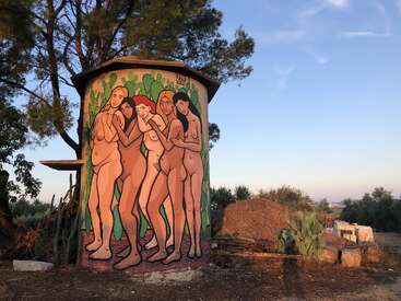 Dieses Bild zeigt eine zylindrische Konstruktion im Freien, die mit einem Wandgemälde verziert ist, auf dem fünf nackte Frauen zu sehen sind, die gemeinsam in einer ländlichen Landschaft bei Sonnenuntergang stehen, umgeben von grünen Pflanzen.