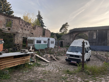 Un vieux bâtiment en pierre délabré entoure une cour où se trouvent une caravane d'époque, une camionnette blanche, des matériaux de construction éparpillés et de l'herbe haute, le tout sous un ciel crépusculaire.