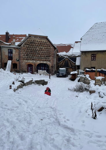 Une cour enneigée bordée de maisons rustiques en pierre. Un petit enfant vêtu d’une combinaison de ski rouge joue dans la neige. Des empreintes de pas et le calme hivernal imprègnent la scène.