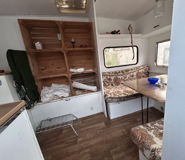 Cette photo montre l'intérieur chaleureux d'un camping-car, avec des banquettes rembourrées à motifs floraux, une table en bois, des étagères ouvertes, une fenêtre et un sol imitation bois. Des couvertures sont rangées.
