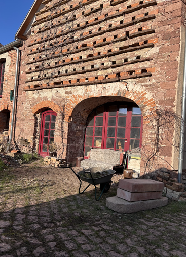 Un bâtiment rustique en briques, avec des portes rouges cintrées, des motifs en treillis et des plantes grimpantes. Une brouette, un banc et des briques sont disposés dans une cour ensoleillée.