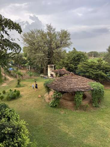 Une scène rurale luxuriante avec de la verdure, un gazebo au toit de chaume, un grand arbre, deux personnes qui marchent, un chien qui se repose et un ciel couvert au-dessus.