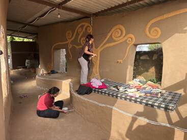 Deux femmes décorent un mur en terre avec des motifs artistiques. Des livres et des couvertures sont éparpillés sur un matelas ; cet espace confortable dégage une atmosphère créative, rustique et accueillante.