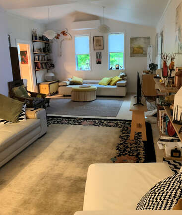 Un salon confortable avec des canapés blancs, des coussins colorés, des étagères, une télévision, des œuvres d'art sur les murs, de grandes fenêtres, des plantes vertes et des éléments décoratifs éclectiques.