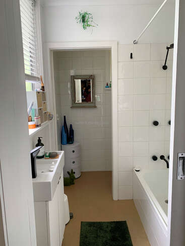 Il s'agit d'une salle de bains claire et bien rangée, avec des carreaux blancs, un petit lavabo, un tapis vert, une baignoire, un miroir, des plantes et divers articles de toilette bien rangés sur les étagères.