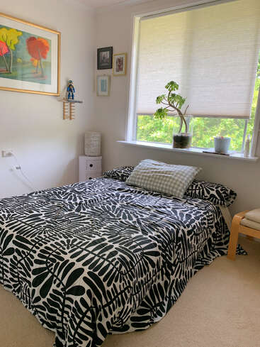 Une chambre à coucher confortable avec un couvre-lit noir et blanc à motifs de feuilles, des murs clairs, des œuvres d'art encadrées, une fenêtre avec des stores, des plantes en pot et un mobilier minimal, créant ainsi une atmosphère calme.