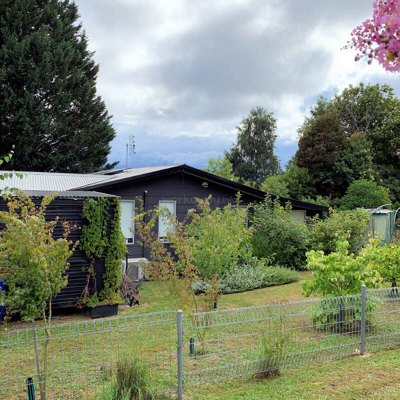 Une petite maison de couleur sombre entourée d'une végétation luxuriante, d'arbres et d'arbustes. Une clôture métallique se trouve devant, sous un ciel nuageux. Cadre rural paisible.