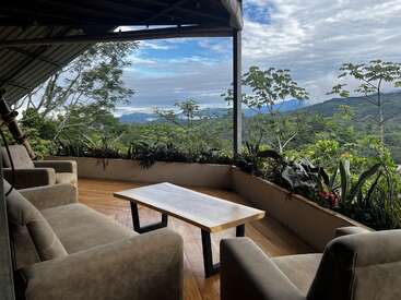 Quatre chaises confortables et une table en bois sont installées sur une terrasse couverte, offrant une vue imprenable sur des montagnes verdoyantes et un ciel lumineux, partiellement nuageux.