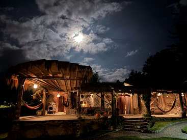 Una cabaña rústica, con una cálida iluminación, se alza bajo un espectacular cielo nocturno, iluminado por una brillante luna llena y nubes dispersas, con hamacas que cuelgan de forma tentadora en el exterior.