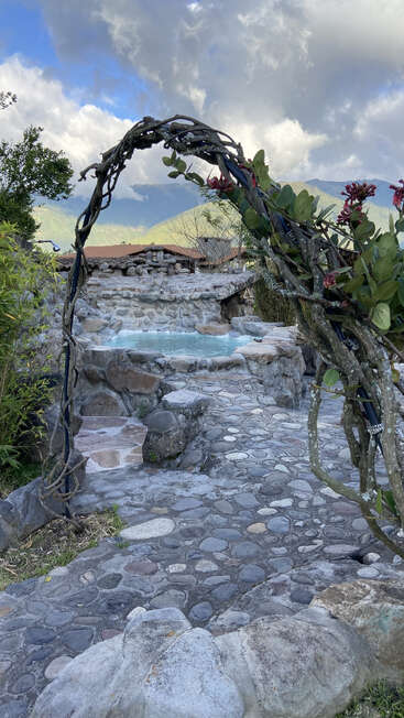 Un sendero de piedra conduce, bajo un rústico arco de parras, a una piscina termal de aspecto natural, rodeada de vegetación, montañas y espectaculares nubes al fondo.